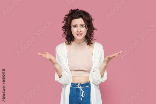 Portrait of angry attractive young adult woman with curly hair wearing casual style outfit raised her hands, asking what, arguing, frowning face. Indoor studio shot isolated on pink background.