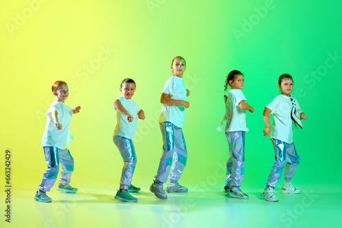Cool girls in fashion outfit with bright glittered makeup performing new dance tricks, synchronous movements over gradient yellow-green background.