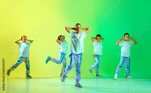 Group of little cute girls wearing style clothes with trendy, creative hairstyle hold hands in choreography class against studio background in yellow-green neon light.