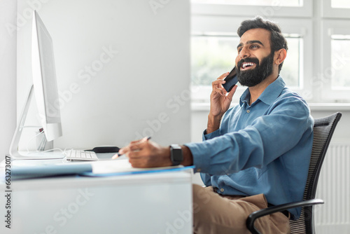 Positive thoughtful indian businessman talking on cellphone and taking notes while working on computer in office