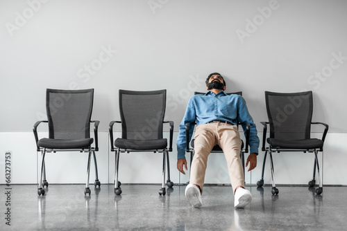 Bored and disappointed male indian employee sitting in waiting room on row of chairs, full length shot