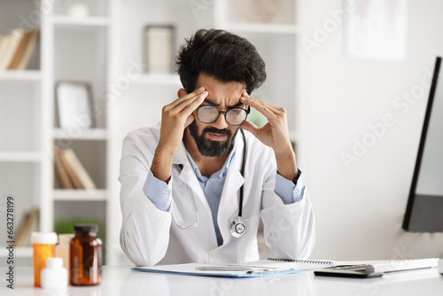 Exhausted hindu man doctor sitting at desk, touching head