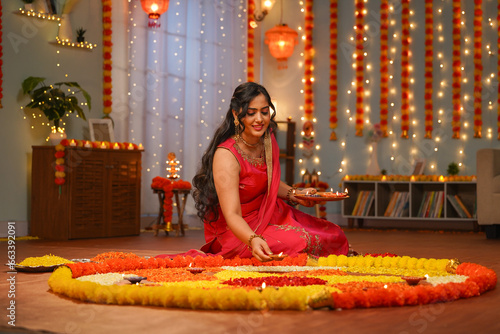 Happy young Indian woman placing diya lamps on decorated flower rangoli for diwali festival celebration at home - concept of festive preparation, traditional culture and spirituality