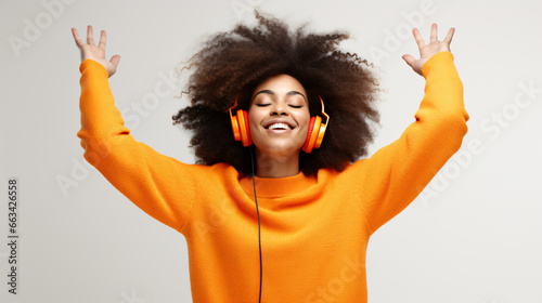 Happy young Afro American woman carried away with music dances carefree with arms raised keeps eyes closed wears stereo headphones on ears dressed in orange jumper isolated over white background