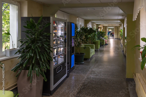 Side view of free standing coffee and combo food (snack, cold drink and food) vending machines standing in corridor with green sofas and plants in public clinic. Copy space for your text.