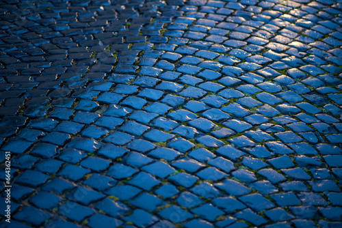 Cobblestone street in Iserlohn Sauerland Germany. Wet shiny historic basalt ashlars or blocks reflecting blue sky and flash light of a car after rainy night. Old pavement background with structures.