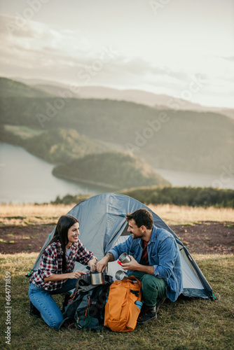 Adventurous couple enjoys a serene moment while camping on a picturesque hilltop overlooking a tranquil lake in the distance