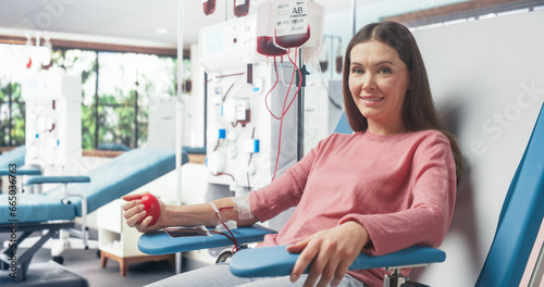 Caucasian Woman Donating Blood For People In Need In Hospital. Female Donor Squeezing Heart Shaped Ball To Pump Blood. Looking Into Camera And Smiling. Donation Center For Victims Of Accidents.