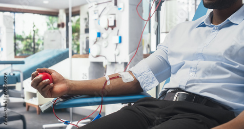 Close Up Shot Of Hand Of Black Male Blood Donor With an Attached Catheter. African Businessman Squeezing Heart-Shaped Ball To Pump Blood Through Tubing Into Bag. Donation For Organ Transplant Patients