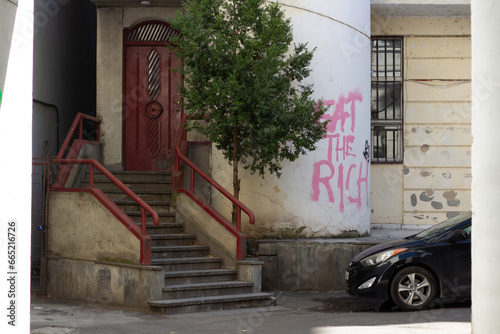 Tbilisi, Georgia - October 2, 2023: House with porch. White wall with text eat the rich. Car parking. Georgian courtyard. Train house with tree. Concept of rent apartment, mortgage, real estate agency