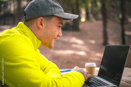 Beautiful caucasian boy influencer working on laptop sitting outdoors in the forest. Hipster young man traveler working distantly while enjoying nature landscape during vacations