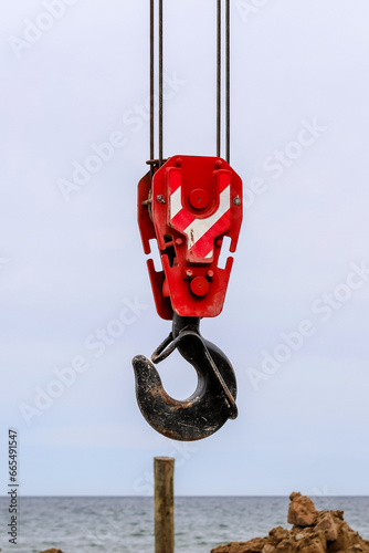 An old lifting hook attached to sheave suspended by wire rope from boom of crane. Construction work on the seashore