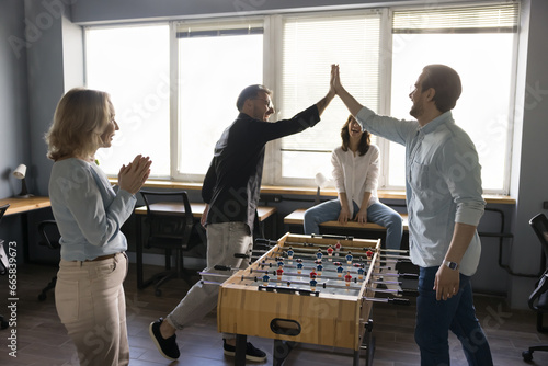 Group of businesspeople rest at lunch break play table foosball, have fun at workplace, enjoy teambuilding activity. Two young man colleagues give high five over board game celebrate final match over