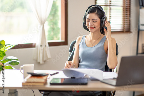 Freelancer Young business asian woman wear wireless headphone and working at home with laptop and papers on desk