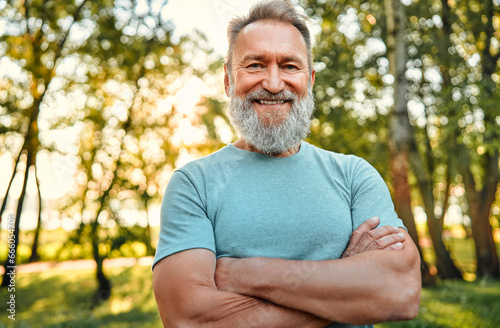 Vitality and active lifestyle. Portrait of fit mature man with hands crossed posing on camera during active training in green forest. Grey-bearded sportsman in blue shirt making confident smile.