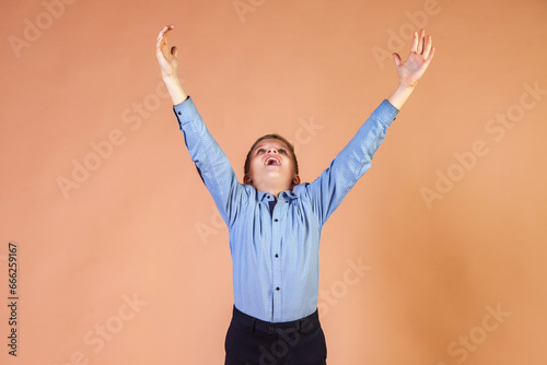 Joyful school boy 11 year old in blue shirt arms raised at beige background, scream looking up. Guy model showing positive emotion, im happy. Children emotions, education concept. Copy ad text space
