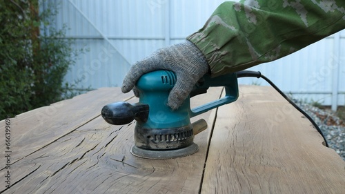 carpenter in gloves works with a sander in a green case with a round emery attachment on polishing solid wooden oak boards in an outdoor workshop, smoothing the surface of the wood with emery