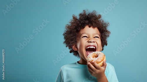 A little boy with a smile is eating a donut on a blue background wall. The child is having a good time with the donut. It's a fun time to have sweet food at home.