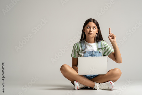 Clever teenage girl sitting on floor with laptop having great idea, finding inspiration, solution to problem. Positive schoolgirl pointing finger up over grey studio background thinking of genius idea