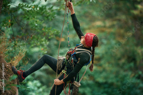 In sportive clothes, high up on the rope. Woman is doing climbing in the forest by the use of safety equipment