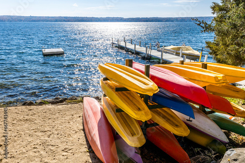 Multi-colored kayaks in a rack on a beach at a lake with a pier and swimming raft on a sunny day.