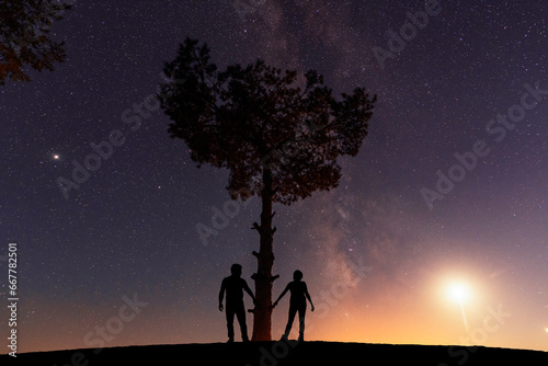 Couple in love standing on the hill near big tree, and looking at the Milky Way galaxy .
