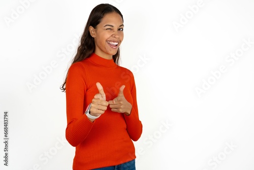 Joyful Beautiful girl wearing red sweater wink and points index fingers at camera, chooses someone, makes finger gun pistol.