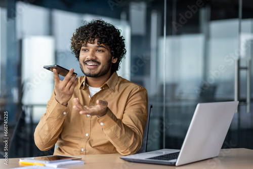 Cheerful and smiling successful businessman using app on phone to record audio message, Indian man inside office with laptop working. A financier talks on the phone, uses an assistant.