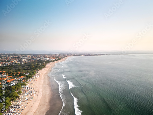 Aerial view of Beautiful sunset on the double six beach with Hotel, resort, cafe buildings, beach umbrella and calm ocean wave in Bali. Tourists are surfing, swimming and walking along the beach.