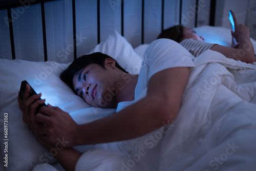 Asian insomnia couple boy and girl addicted playing social media and texting message to friend with blue light from mobile screen when turned their backs sleep in bed at the dark night bedroom