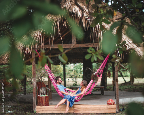 casal jovem deitado em rede cor de rosa em frente a uma maloca com teto de palha rodeado de árvores verdes, em Soure, ilha do Marajó 