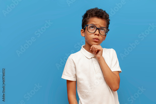 pensive african american teenager boy in glasses thinks and plans on blue isolated background, curly schoolboy imagines