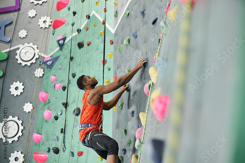 young african american man climbing up bouldering wall with safety rope and alpine harness
