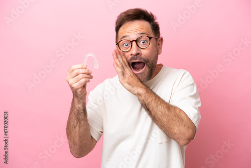 Middle age man holding invisible braces isolated on pink background whispering something