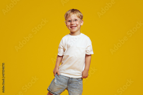 Little student boy in stylish white tshirt and eyeglasses smiling and looking at camera. Cute child laughing and standing isolated over yellow background