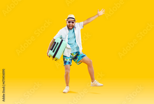 Full length portrait of a funny happy young man in sunglasses and beach summer clothes standing with suitcase isolated on a studio yellow background. Vacation trip and travel concept.