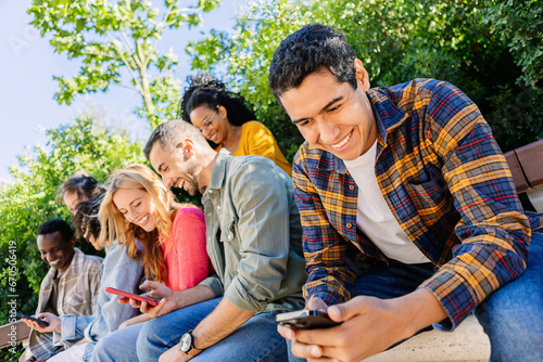 Young group of people using cell phones sitting together outside. Technology lifestyle concept with college student friends enjoying social media content on mobile phone network app.