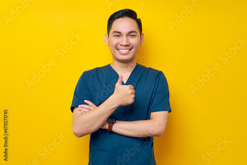 Professional young Asian male doctor or nurse wearing a blue uniform showing thumb up or like, looking at camera with smile isolated on yellow background. Healthcare medicine concept