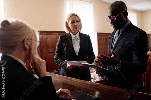 Defense and prosecution sides of court case standing in front of mature female judge while African American attorney explaining some points