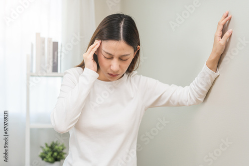 Dizzy asian young woman, girl headache or migraine pain. suffering from vertigo, difficulty standing up leaning on wall at home, holding head with hand, health problem of brain, inner ear not balance.