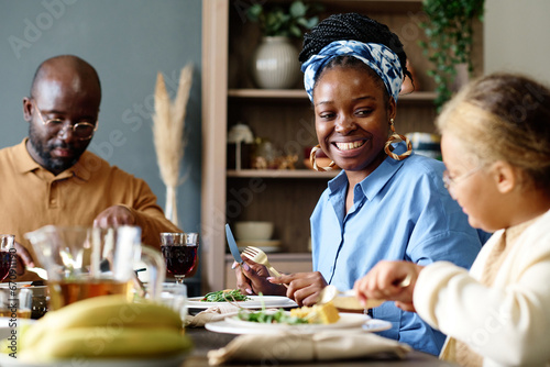 Focus on happy young African American woman looking at her daughter by festive table while sitting between her husband and cute girl