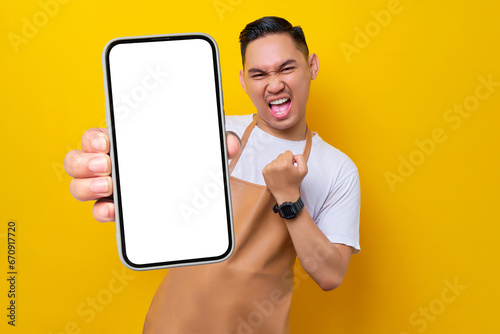 excited young Asian man barista barman employee wearing brown apron work in coffee shop holding mobile phone and showing winner gesture isolated on yellow background. Small business startup concept