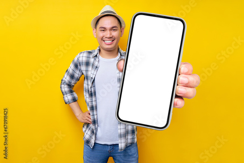 Cheerful handsome Asian young man 20s wearing casual clothes hat standing while hand on his waist, Showing mobile cell phone with blank screen isolated on yellow background. People lifestyle concept