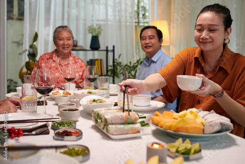 Smiling woman taking spring roll and bowl of sauce at family dinner