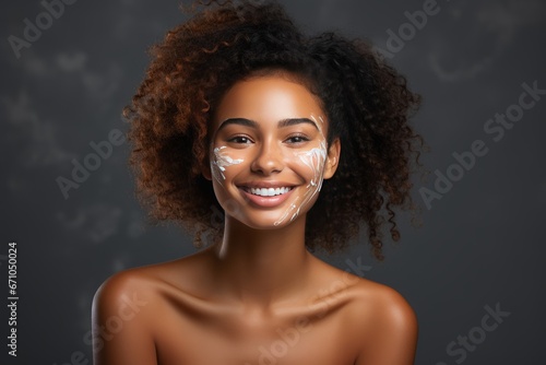 Close-up of young African American woman with moisturizer on her face. Smiling face of beautiful colored lady with daily cream, facial cosmetics. Skin care. Monochrome background, copy space.