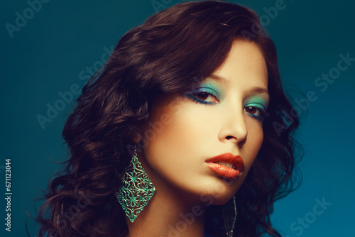 Epoque of Disco concept. Close up portrait of fashion model with curly hair and arty make-up posing over blue background. Vintage turquoise earrings. Perfect skin. Retro style. Copy-space. Studio shot