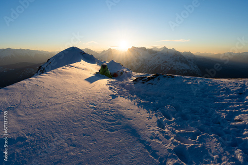 Winterbiwak im Schnee auf einem Berg in den Alpen mit grünem Zelt zum Sonnenaufgang und Blick auf das Wettersteinmassiv und Zugspitze