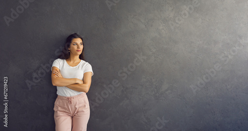 Banner background with confident attractive young woman in casual outfit standing with her arms crossed leaning on dark grey copy space wall, looking away and thinking about something