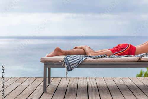 Les jambes d'un homme en maillot de bain sur transat en terrasse, serviette pend du transat, mer et ciel en arrière-plan. Instant de détente au bord de l'eau
