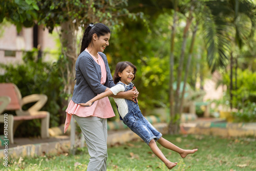 focus on mother, Cheerful indian mother playing by rotating his daughter at park - concept of weekend holidays, Carefree moments and Playful affection.